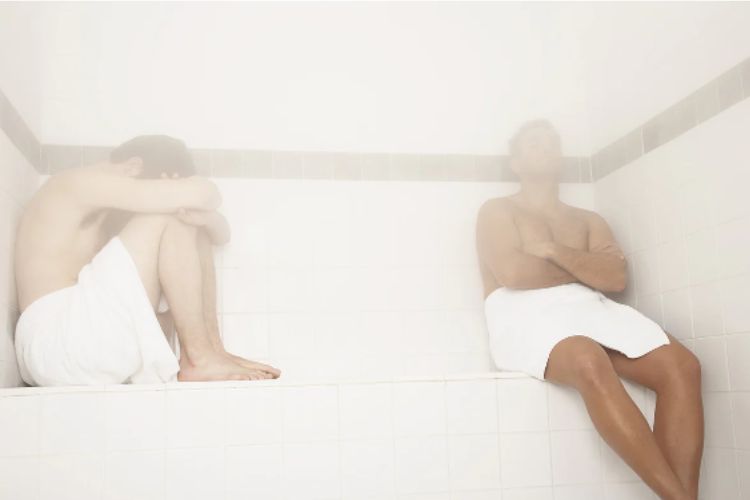 Two individuals in a steamy sauna room, one seated with head down and the other leaning against the wall, both wearing towels around their waists.